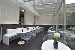 A modern outdoor seating area with white benches and round tables, some with vases of flowers.