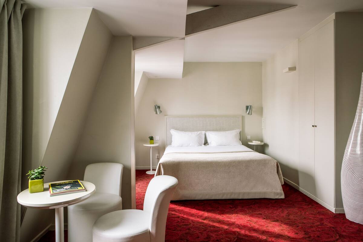 A hotel room with a bed, a small table with a plant and a book, a chair, and a red carpet.