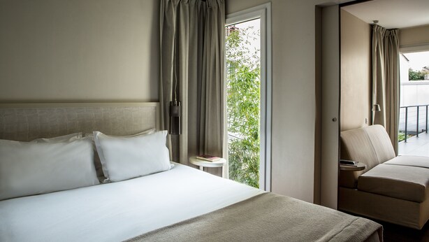 A hotel room with a bed, a chair, a small table, and a view of greenery outside. - Hôtel Le Quartier Bercy Square (Paris)