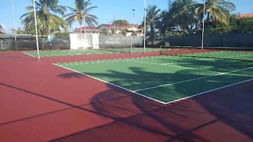 Ein Tennisplatz mit roter Belag und grünen Linien, umgeben von Palmen und einem Gebäude im Hintergrund.