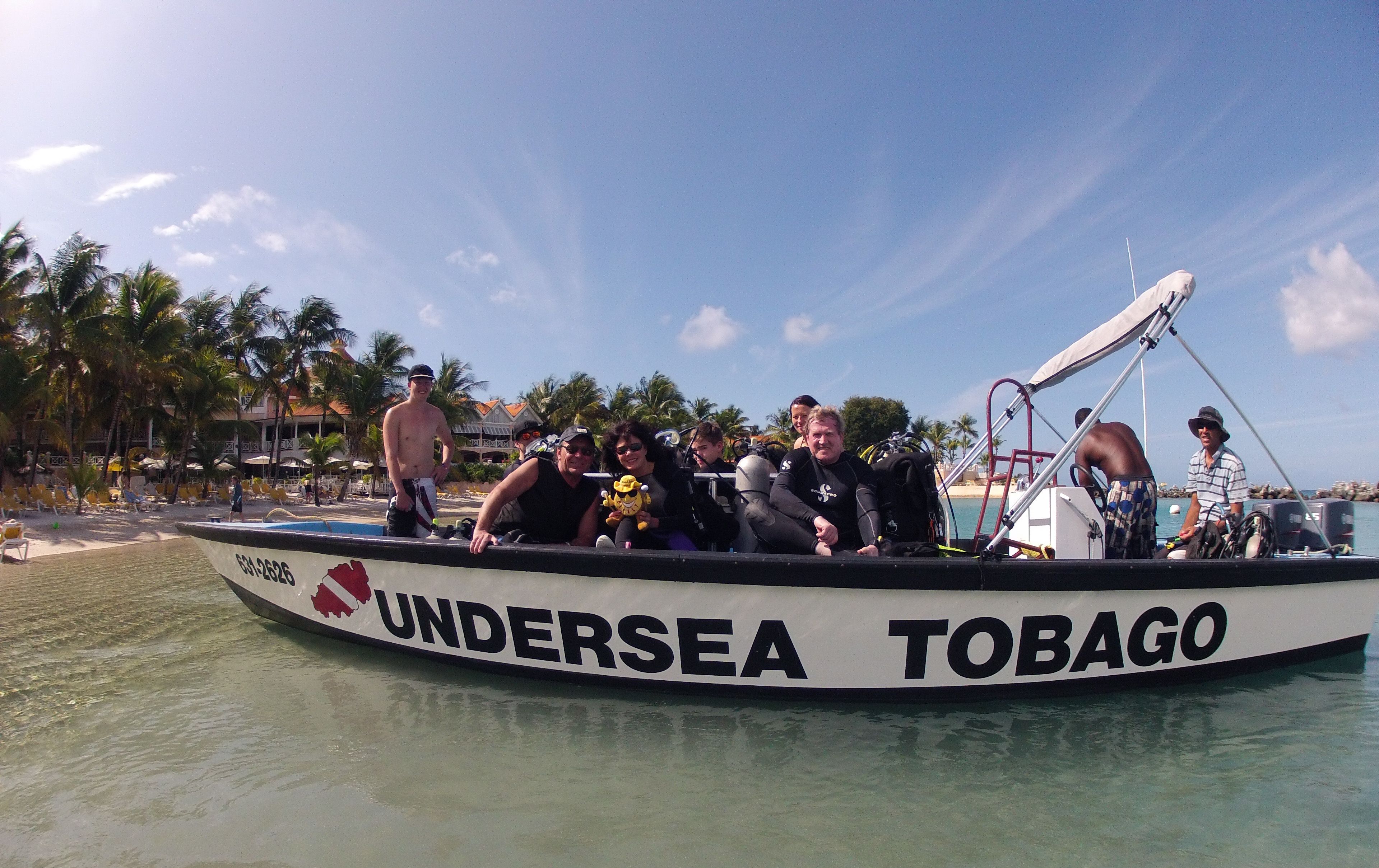 A boat named 'Undersea Tobago' with people on board, a clear sky, and palm trees in the background.