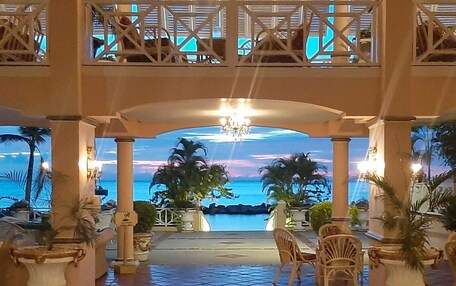 A hotel lobby with a view of the ocean, featuring a tiled floor, wooden chairs, and potted plants.