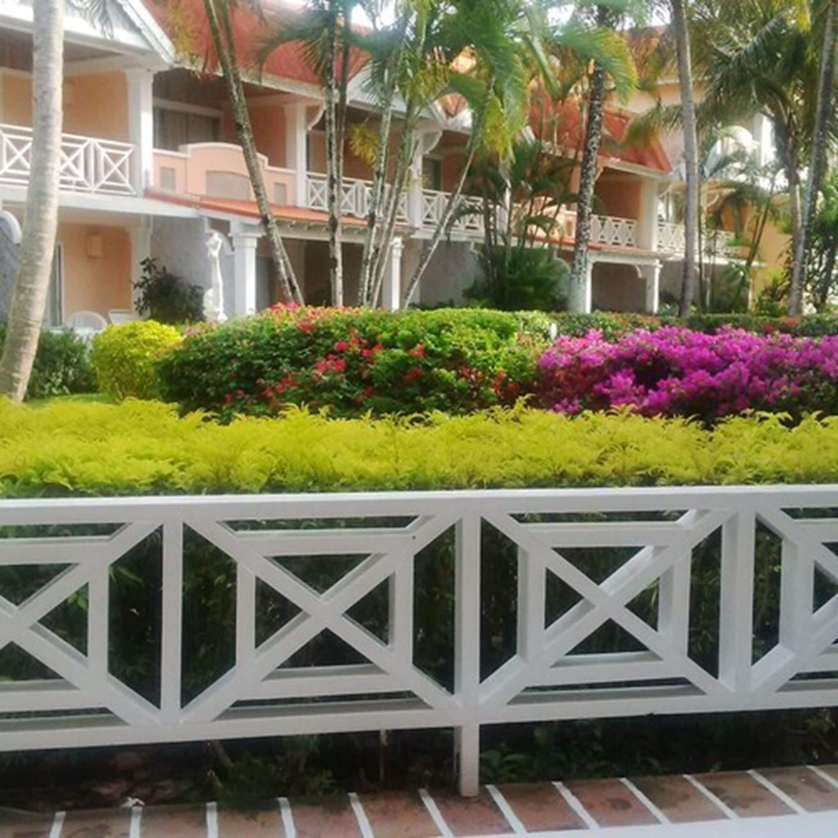 A hotel with a white balcony railing, lush green and pink bushes, and palm trees.
