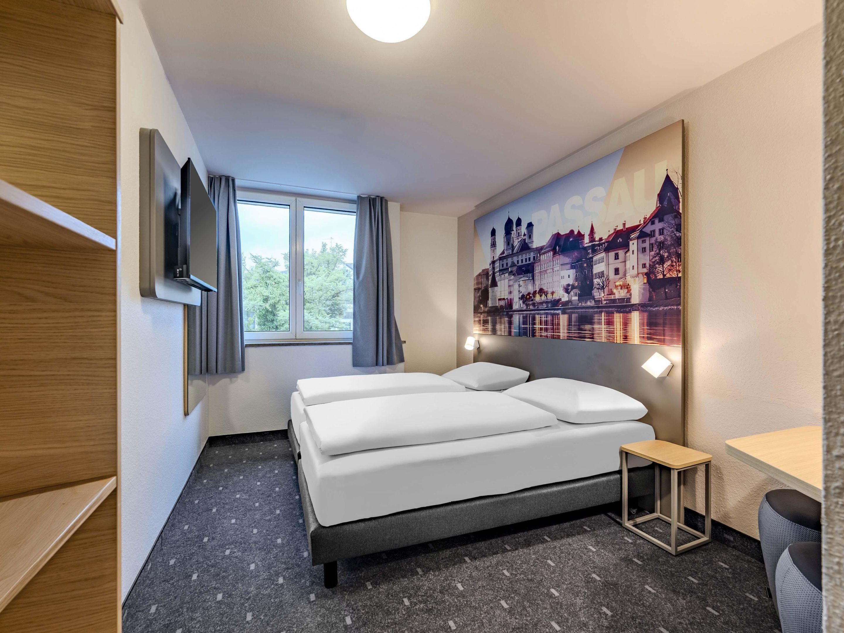 A modern hotel room with a bed, a small table, a TV, and a large window with curtains.