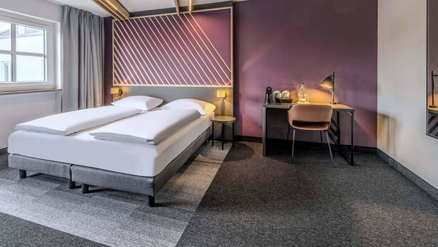 Una habitación de hotel moderna con cama, escritorio, silla y una pared de acento morada.