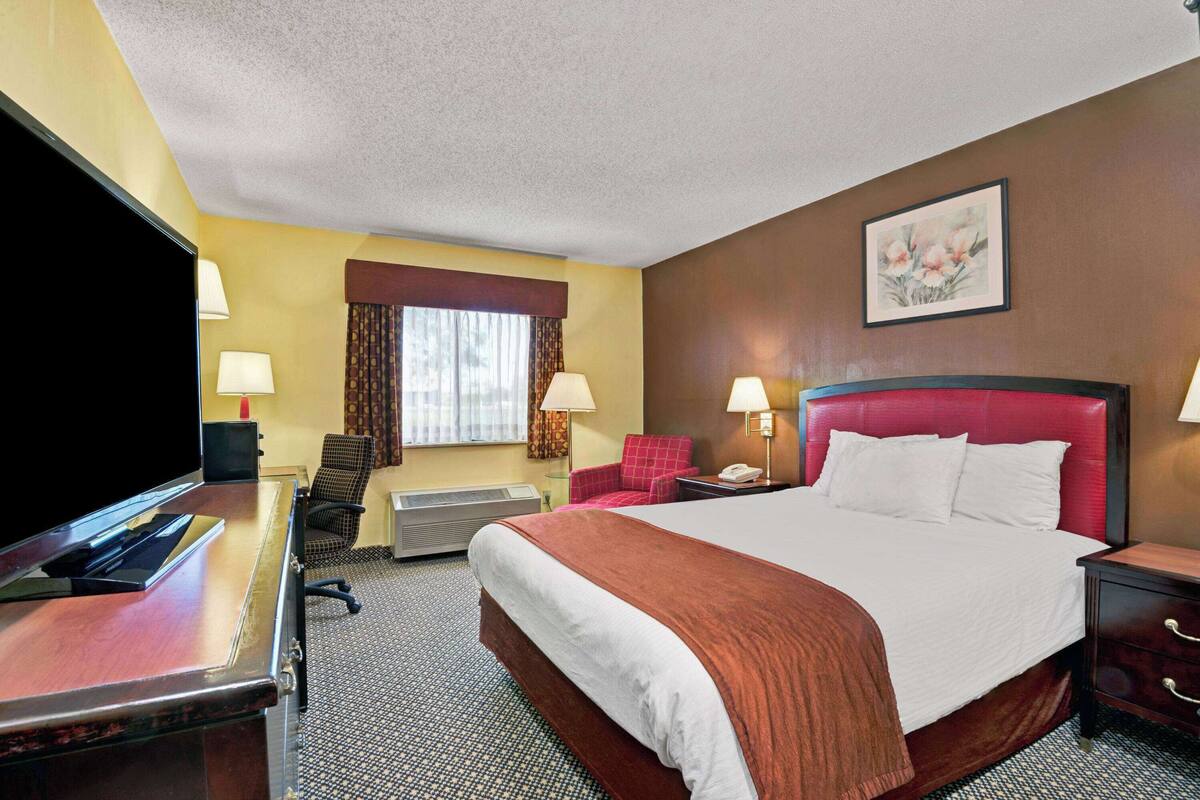 A hotel room with a large bed, a desk with a flat-screen TV, two lamps, a chair, and a painting on the wall.