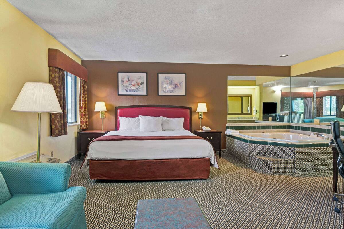 A hotel room with a large bed, a sitting area, a bathroom with a jacuzzi, and a mirror.