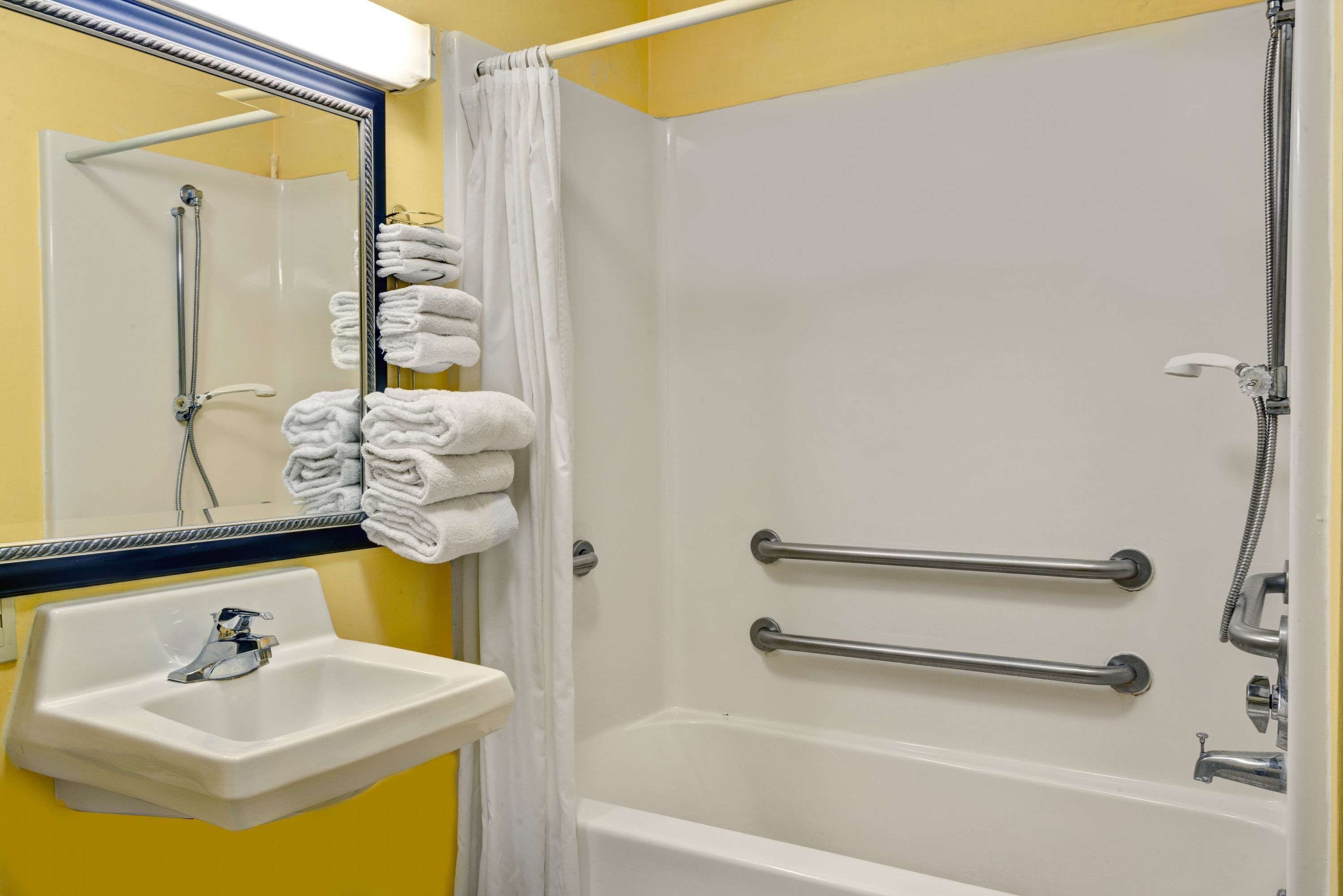 A bathroom with a white sink, a mirror, a shower with grab bars, and neatly stacked towels.