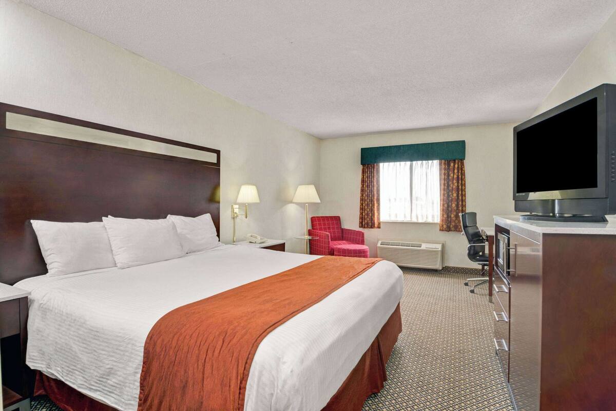 A hotel room with a large bed, a desk with a flat-screen TV, a chair, and a desk lamp.