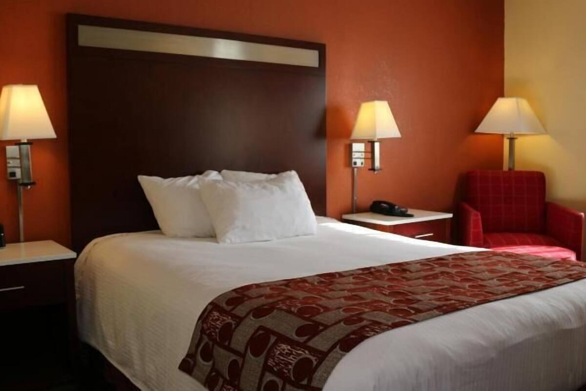 Habitación de hotel con una cama grande, dos mesitas de noche con lámparas, una silla roja y un plaid rojo con estampado.