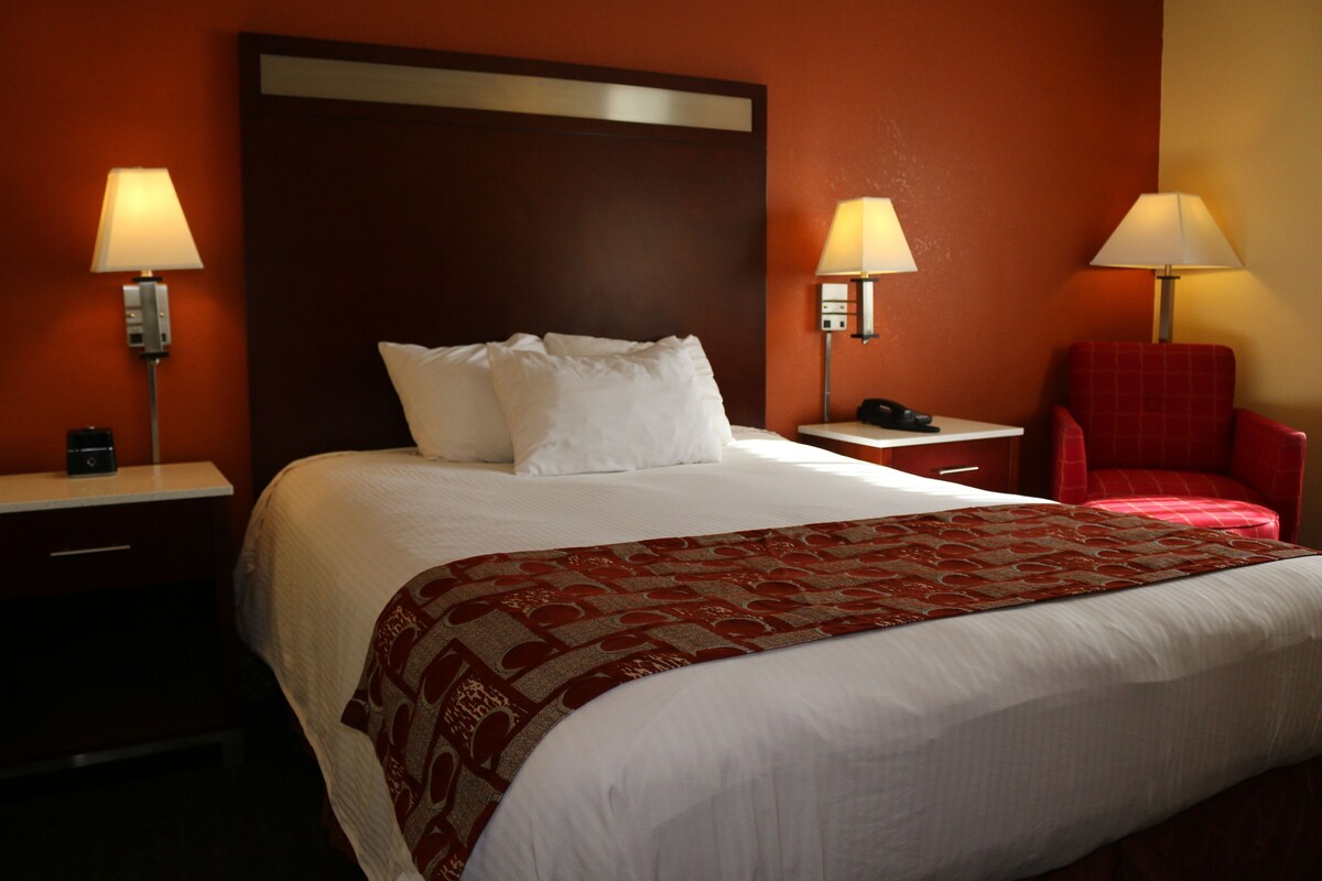 Una habitación de hotel con una cama grande, dos mesitas de noche, una silla roja y dos lámparas de pared.