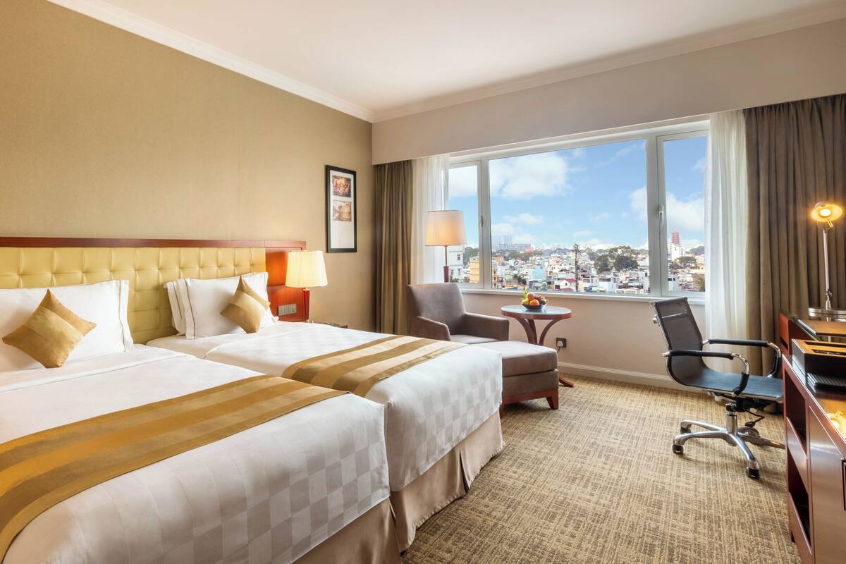 A hotel room with two beds, a desk, a chair, a lamp, and a view of the city.
