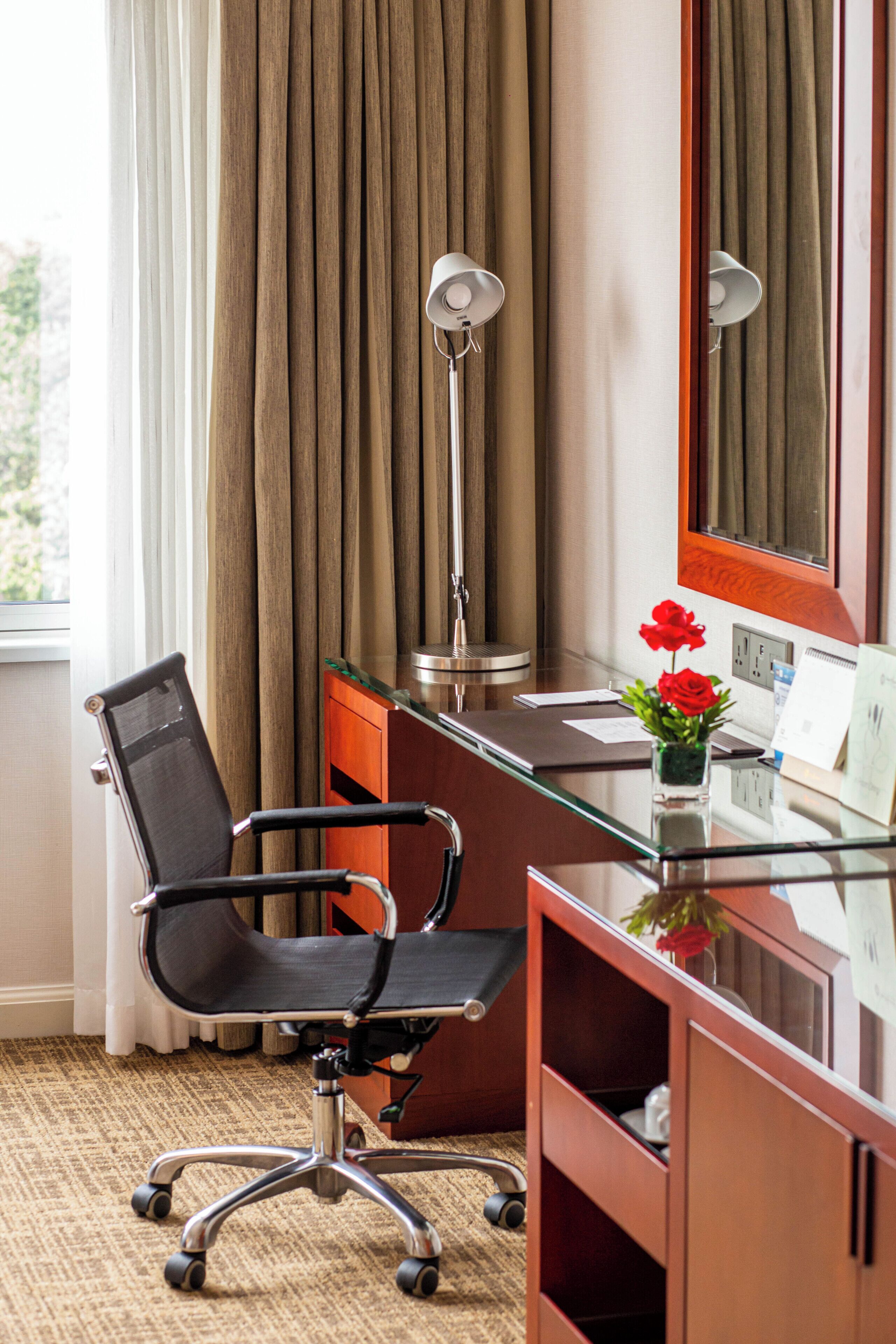 A hotel room with a desk, chair, lamp, and a vase with flowers.