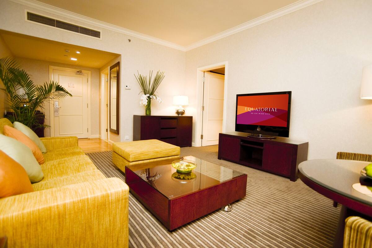 A hotel room with a yellow sofa, a glass coffee table, a flat-screen TV, a lamp, and a potted plant.