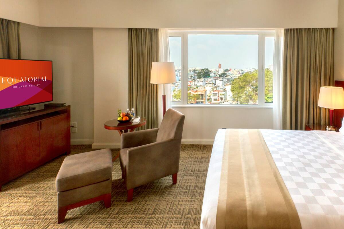 A hotel room with a bed, a TV, a desk, a chair, and a view of the city.