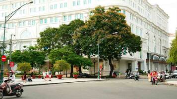 A street view with a large white building, trees, and people walking and riding scooters.