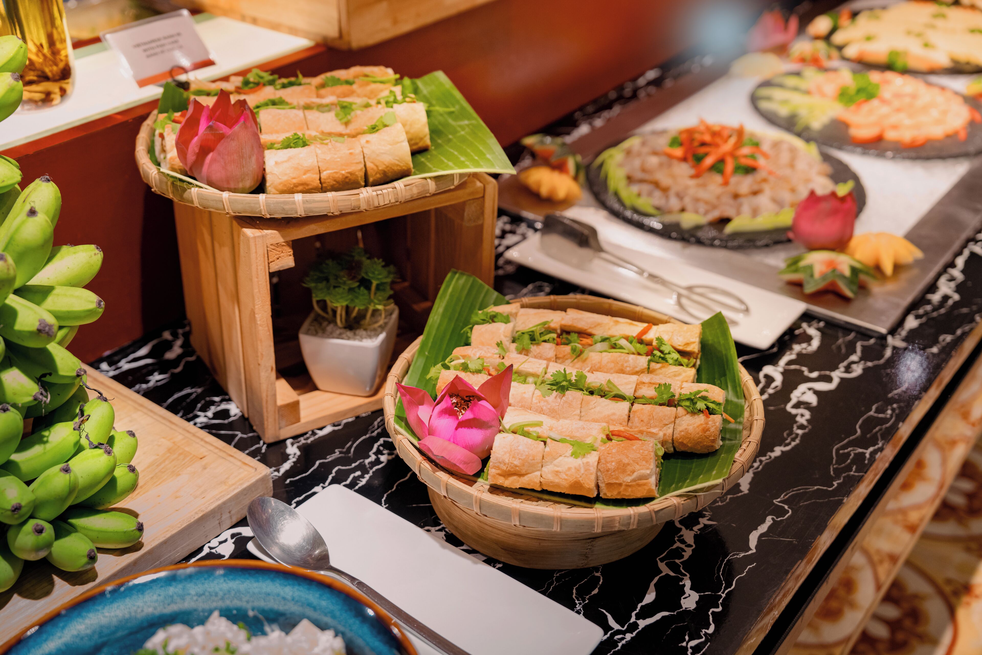 A buffet with various food items, including a dish with cubes on a banana leaf, a plate with rice and vegetables, and a bowl of green bananas.
