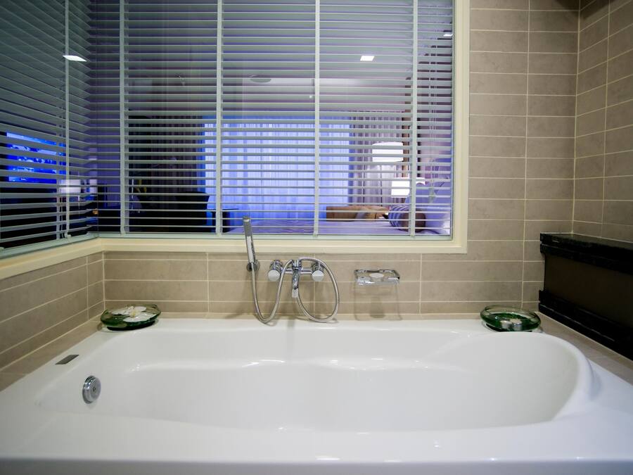 A bathroom with a bathtub, a window with blinds, and a shelf with toiletries.