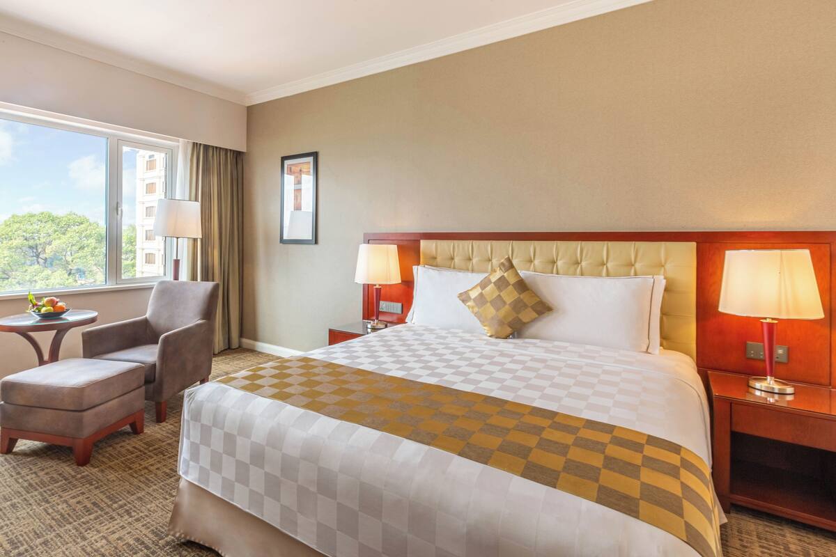 A hotel room with a large bed, two bedside lamps, a chair, a small table, and a window with a view of trees.