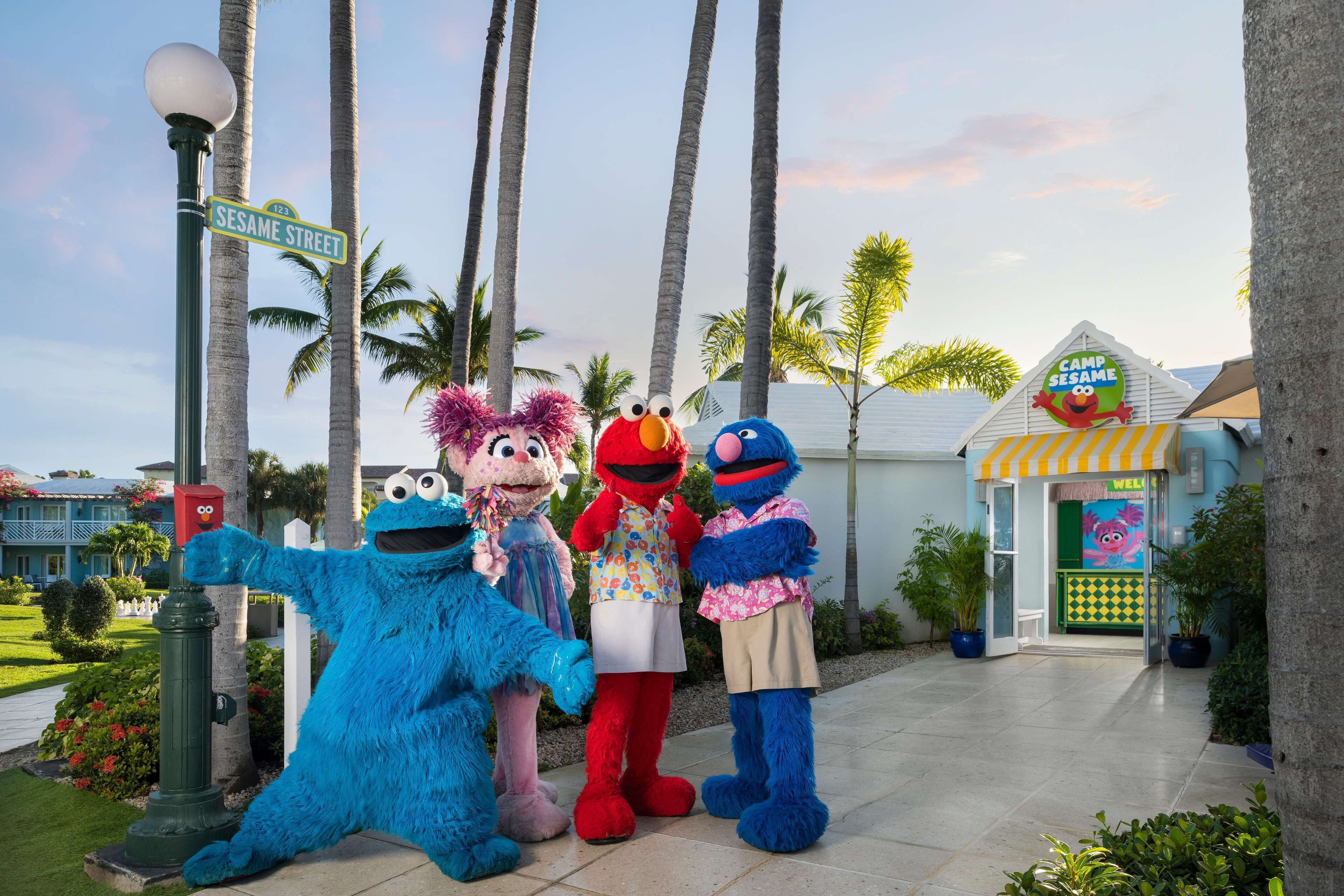 Sesame Street characters standing on a sidewalk with palm trees and a building labeled "Camp Sesame" in the background.