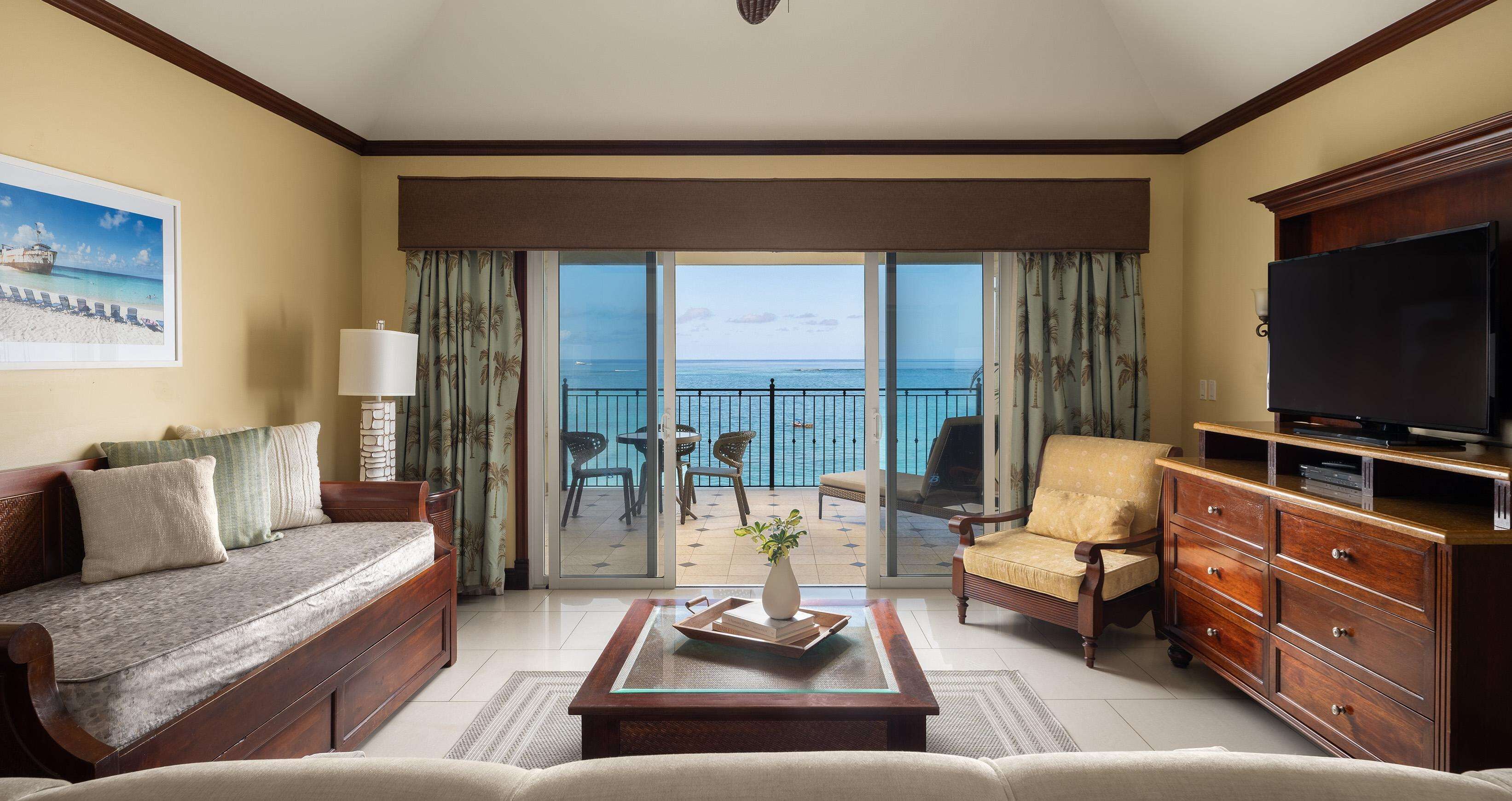 A hotel room with a sofa, armchair, TV, and a balcony with a view of the ocean.