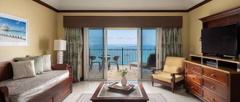 A hotel room with a sofa, armchair, TV, and a balcony with a view of the ocean.