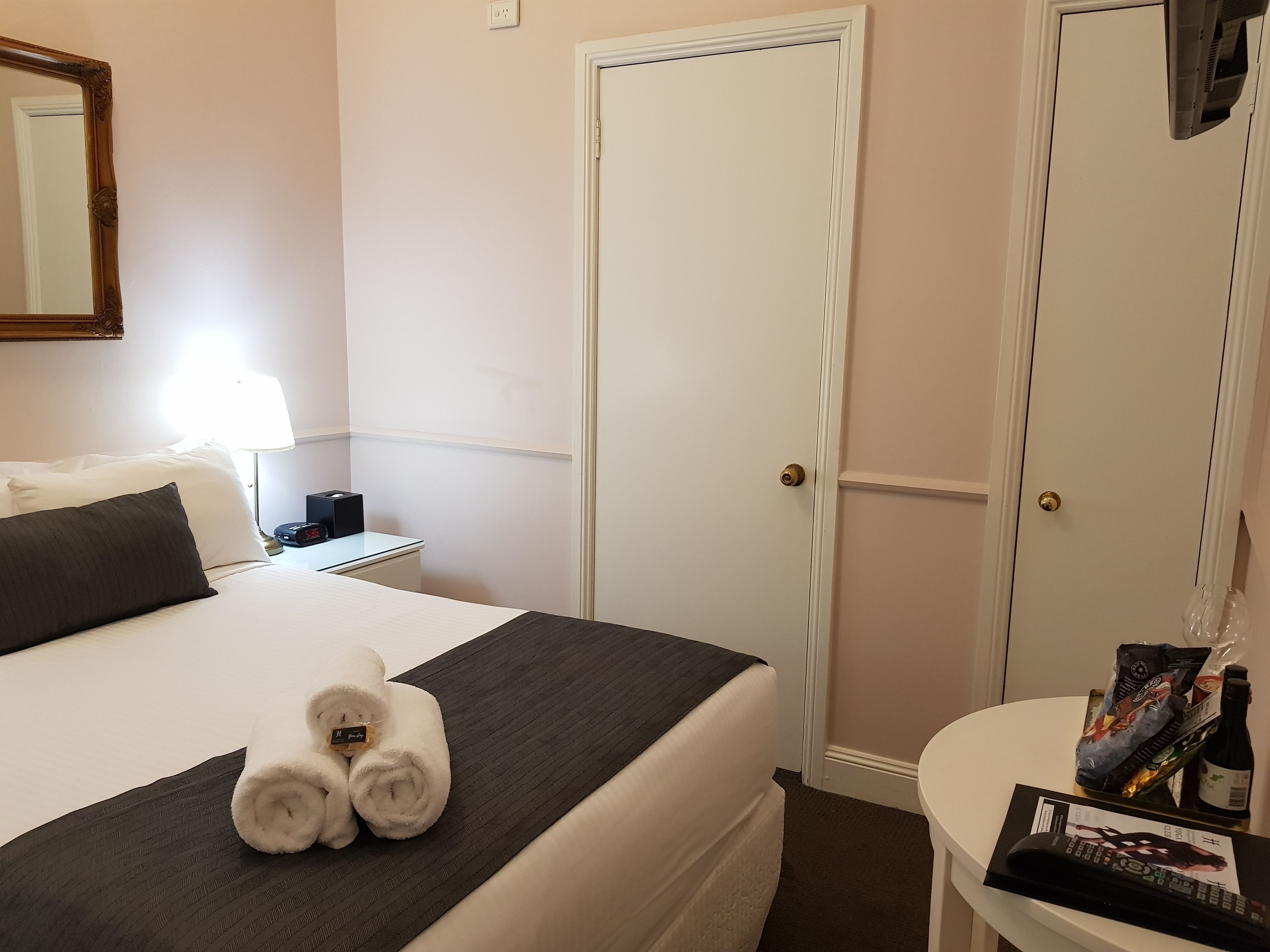 A hotel room with a bed, a bedside table with a lamp, a wall-mounted mirror, a door, and a small table with a remote control and a bottle.