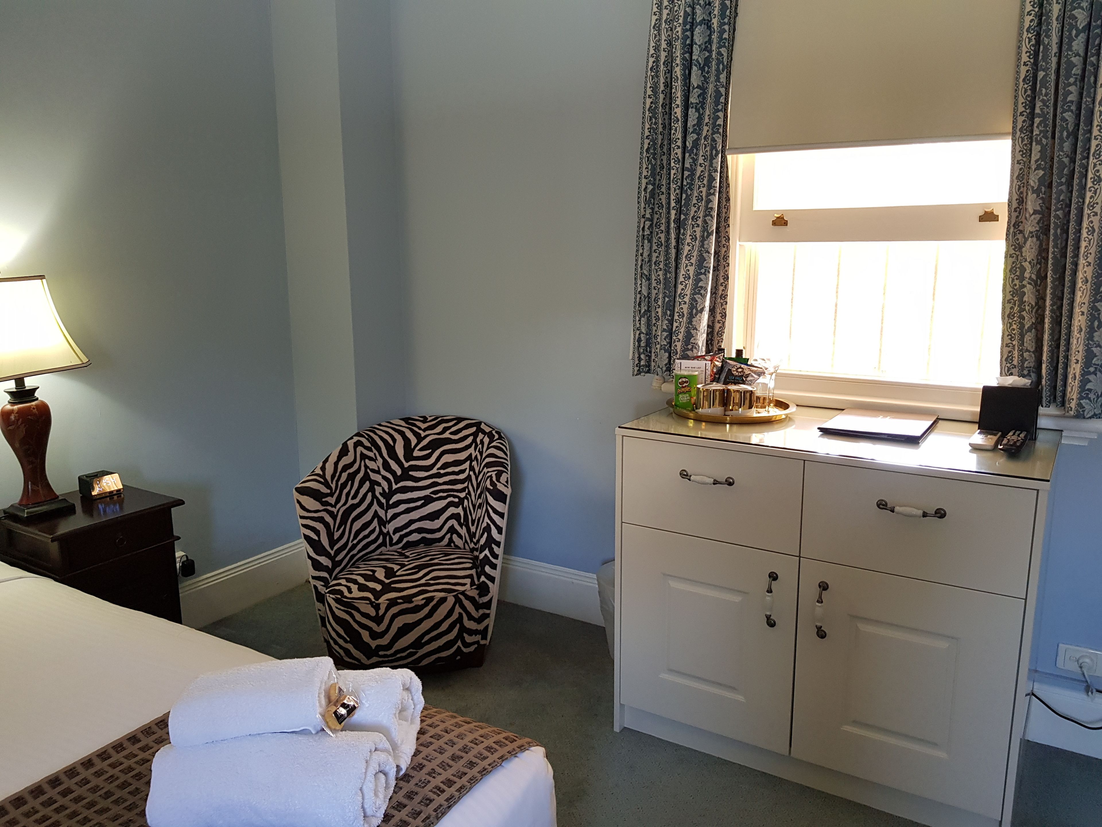 Ein Hotelzimmer mit einem Bett, einem zebra-gemusterten Sessel, einem Schreibtisch mit Lampe und einem fensterbelaubten Fenster.
