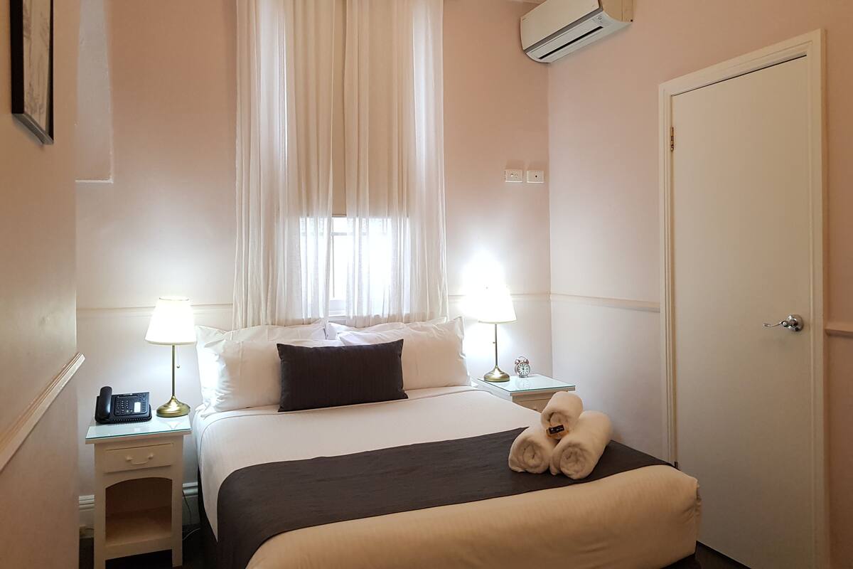 A hotel room with a bed, two bedside lamps, a window with curtains, an air conditioning unit, a bedside table with a phone, and a door.