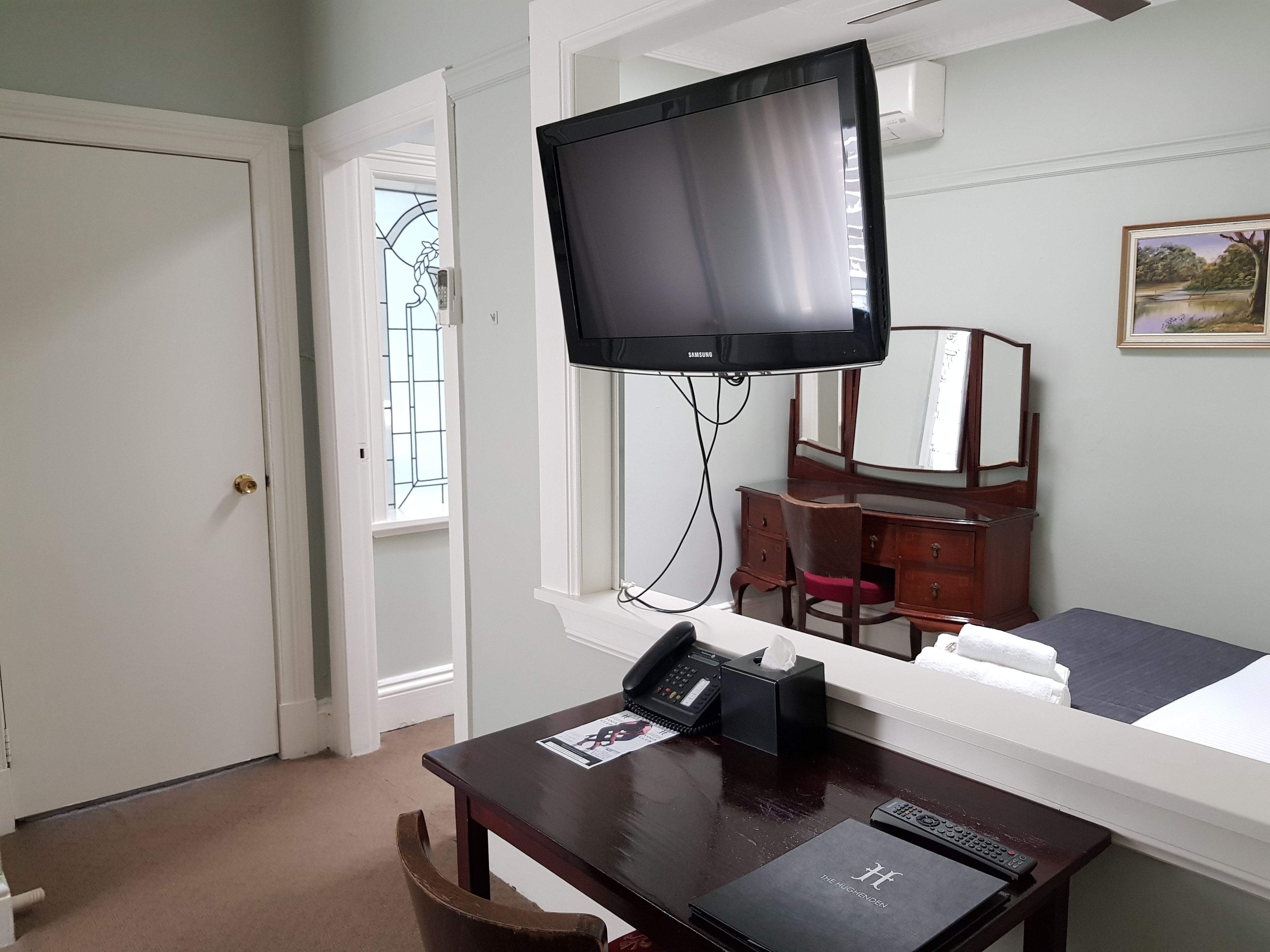 Habitación de hotel con una cama, un escritorio con teléfono, televisor montado en la pared, cómoda y ventana con vista a árboles.