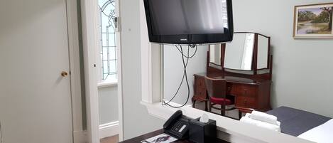 A hotel room with a bed, a desk with a telephone, a television mounted on the wall, a dresser, and a window with a view of trees.