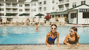 A swimming pool area with lounge chairs and umbrellas, a pool building with a sign, and a multi-story building in the background. - The Osthoff Resort (Elkhart Lake)