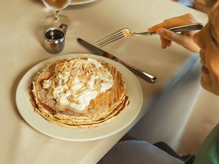 A plate of pancakes with whipped cream and syrup, a fork, and a knife on a table.