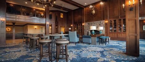 A spacious lobby with a high ceiling, chandeliers, a stone fireplace, and a blue patterned carpet.