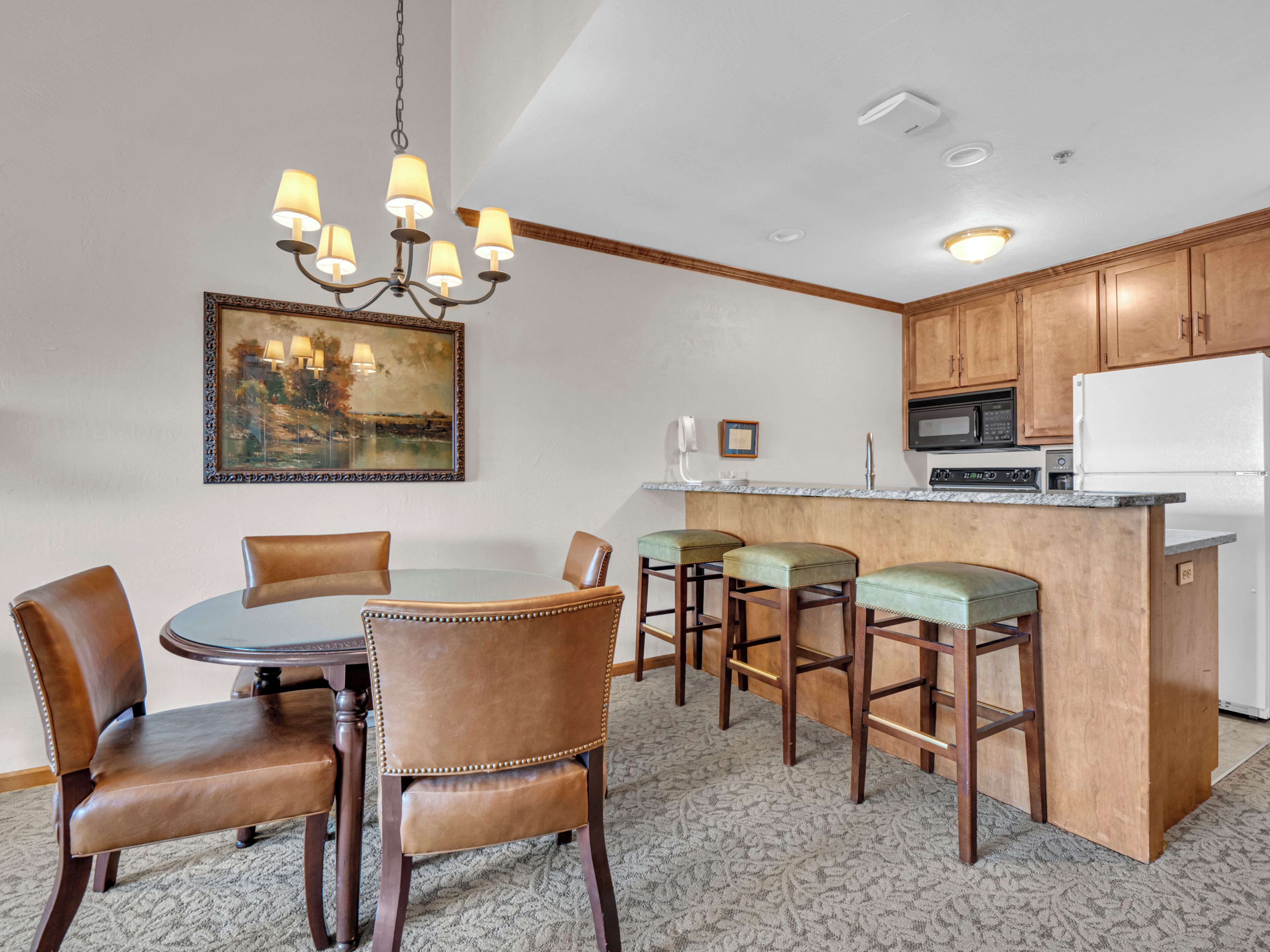 A dining area with a round table, four chairs, a chandelier, and a kitchen with wooden cabinets and appliances.