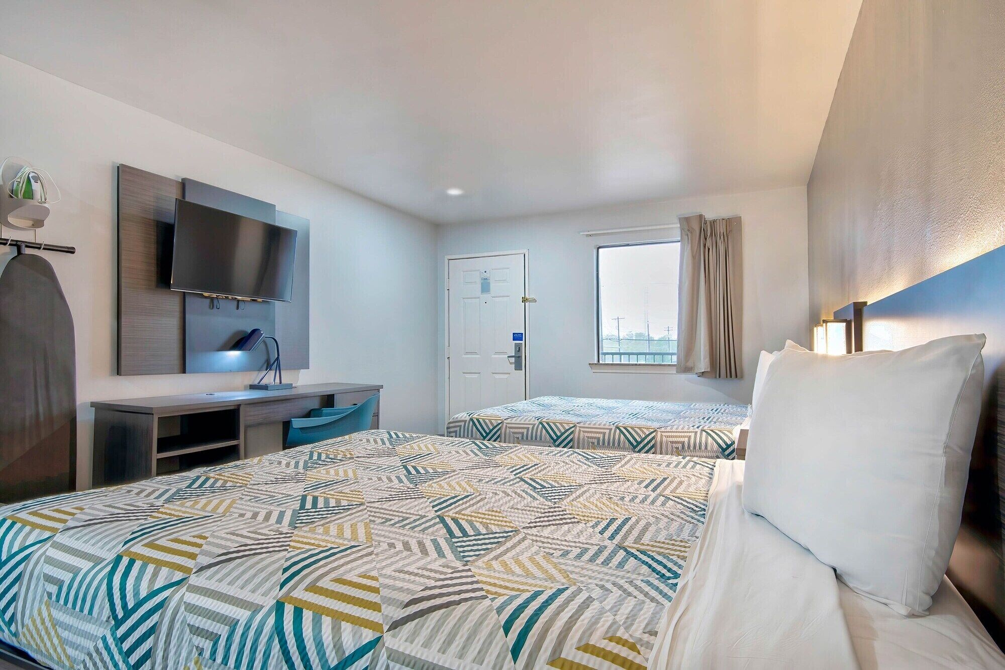 A hotel room with two beds, a desk with a flat-screen TV, a window with curtains, and a door.