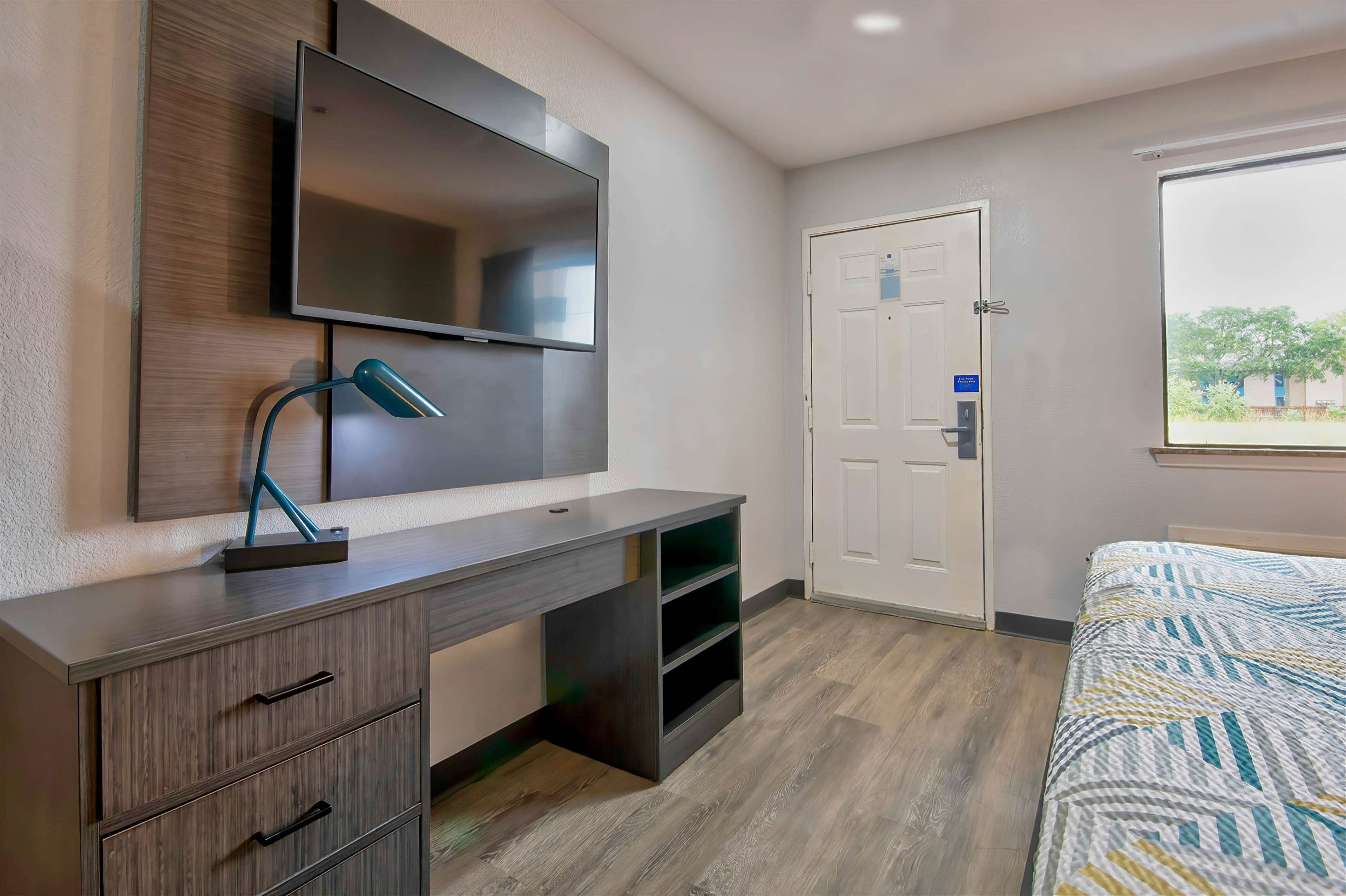 A bedroom with a bed, a desk with drawers, a TV mounted on the wall, and a door with a window.
