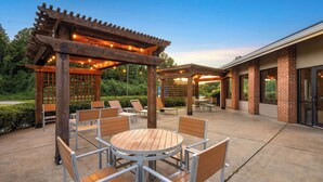 Un espace extérieur aménagé en terrasse, avec des tables et des chaises en bois, une pergola illuminée de guirlandes lumineuses et, au fond, un bâtiment en briques.