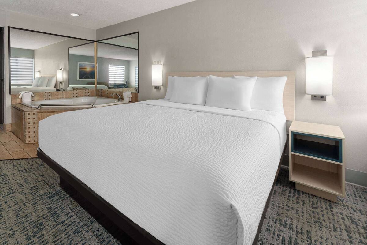 A hotel room with a large bed, a wooden headboard, a nightstand with a lamp, a mirror, and a bathroom with a bathtub and toilet.