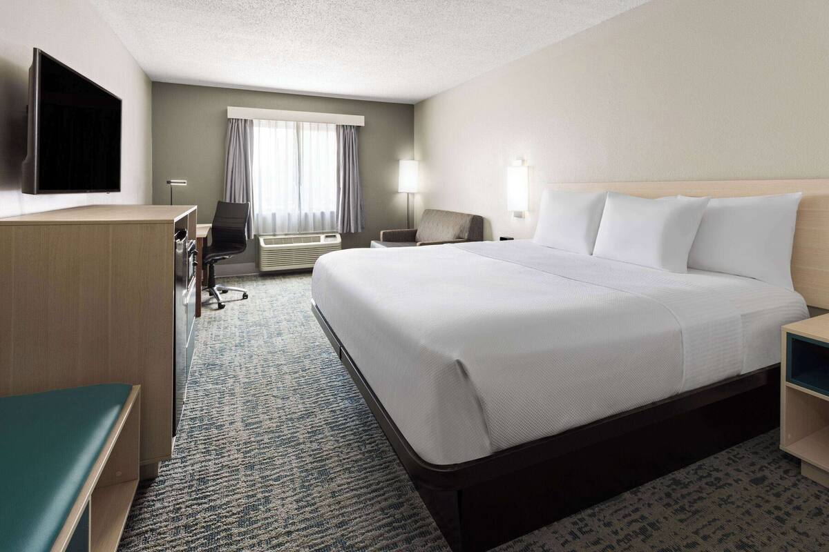 A hotel room with a large bed, a desk with a chair, a flat-screen TV mounted on the wall, and a window with curtains.