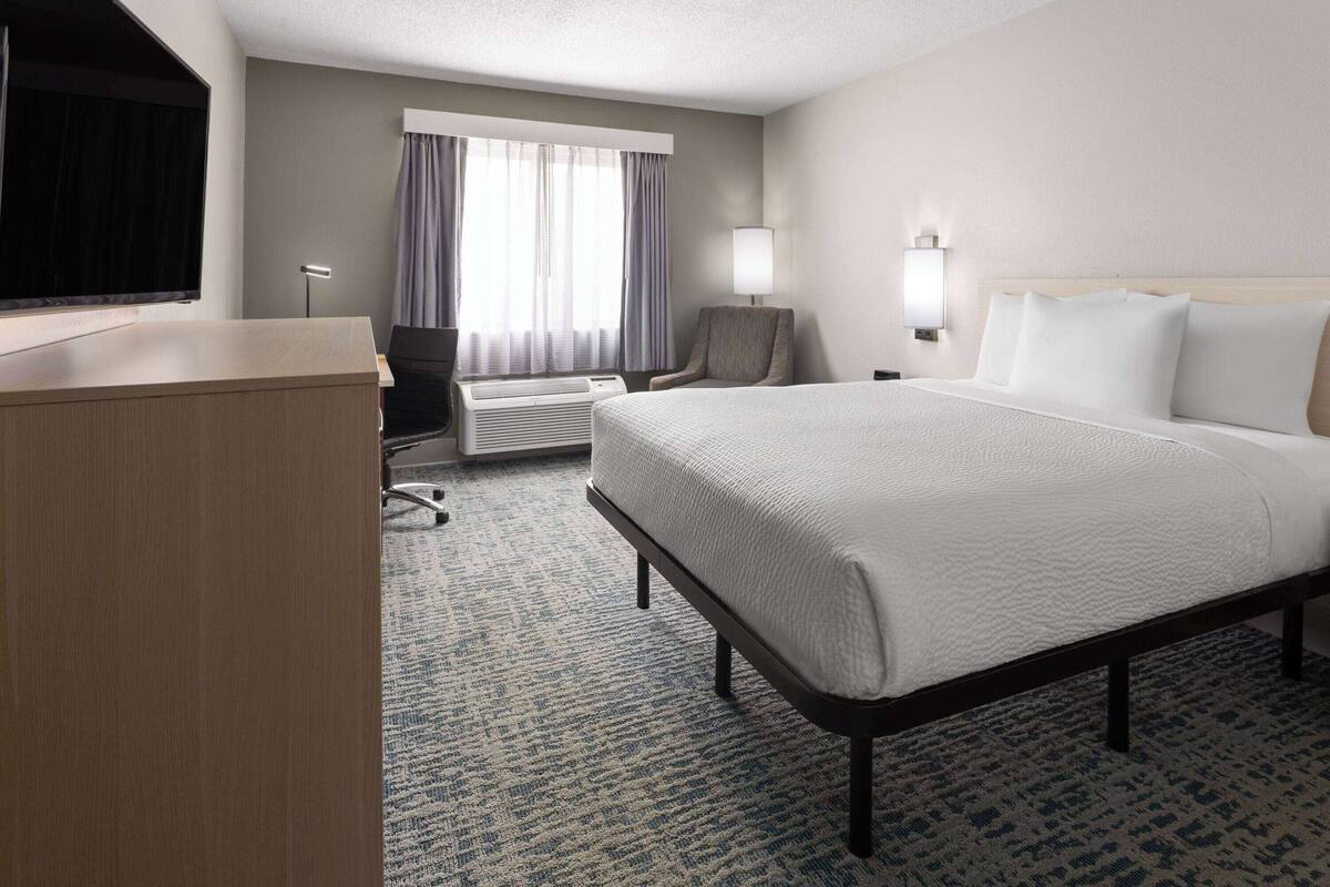 A hotel room with a large bed, a desk, a chair, a flat-screen TV, a window with curtains, and a wall-mounted lamp.
