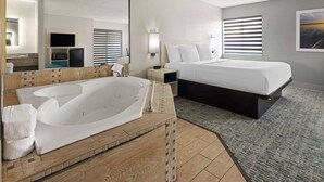 Une chambre d’hôtel dotée d’une grande baignoire, d’un lit avec une literie blanche, d’une coiffeuse en bois, d’un miroir, d’une télévision et d’une fenêtre avec des stores.