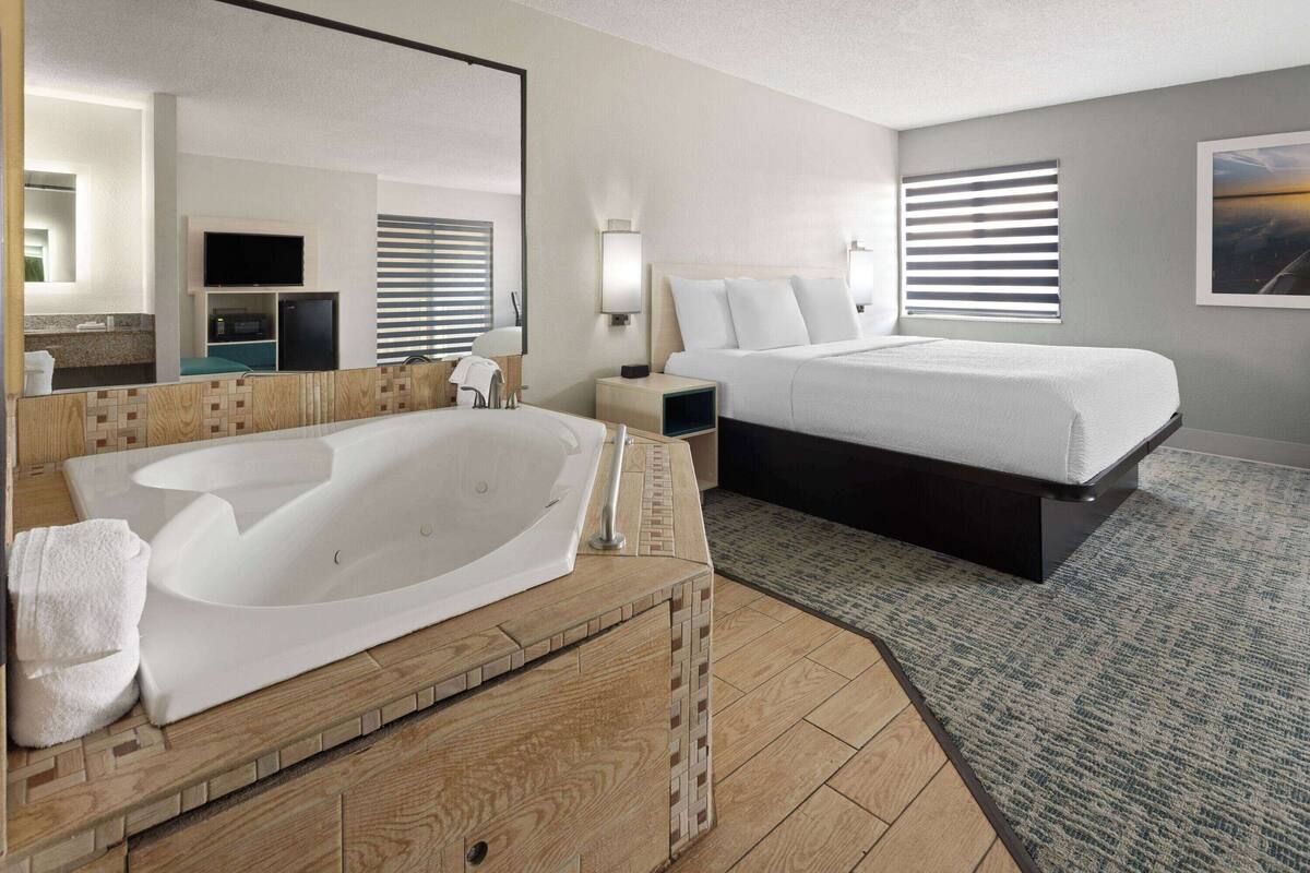 A hotel room with a large bathtub, a bed with white bedding, a wooden vanity, a mirror, a television, and a window with blinds.