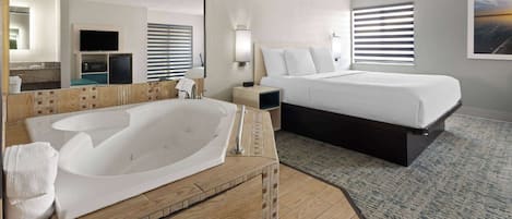 A hotel room with a large bathtub, a bed with white bedding, a wooden vanity, a mirror, a television, and a window with blinds.