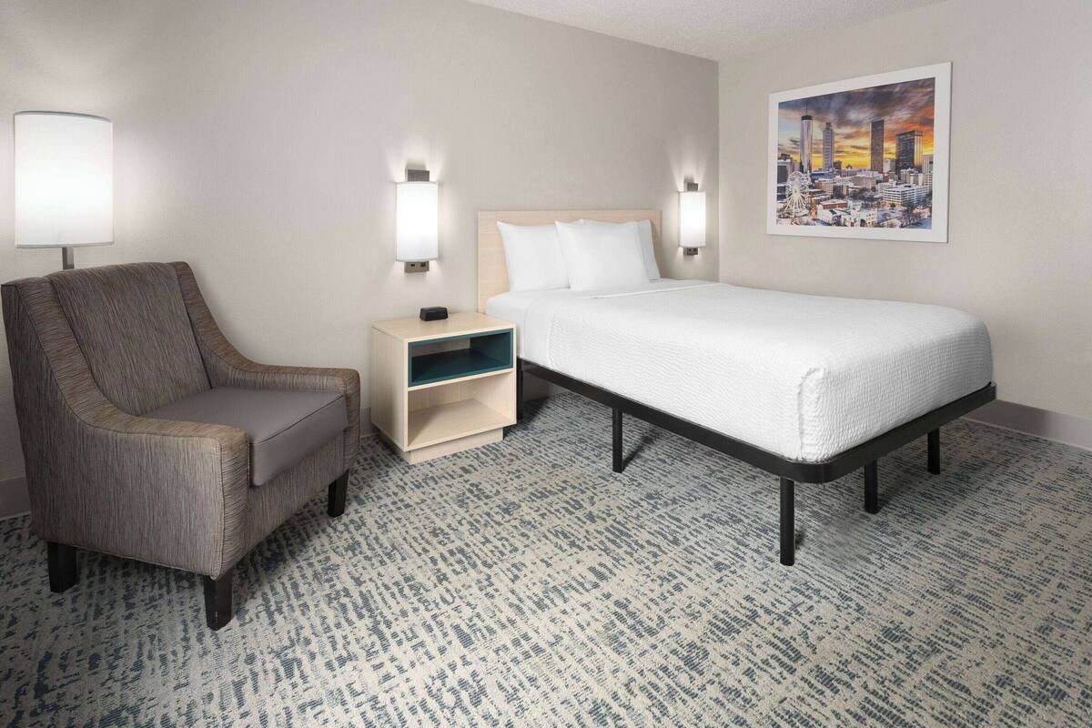 A hotel room with a bed, a chair, a nightstand, and a wall-mounted lamp.
