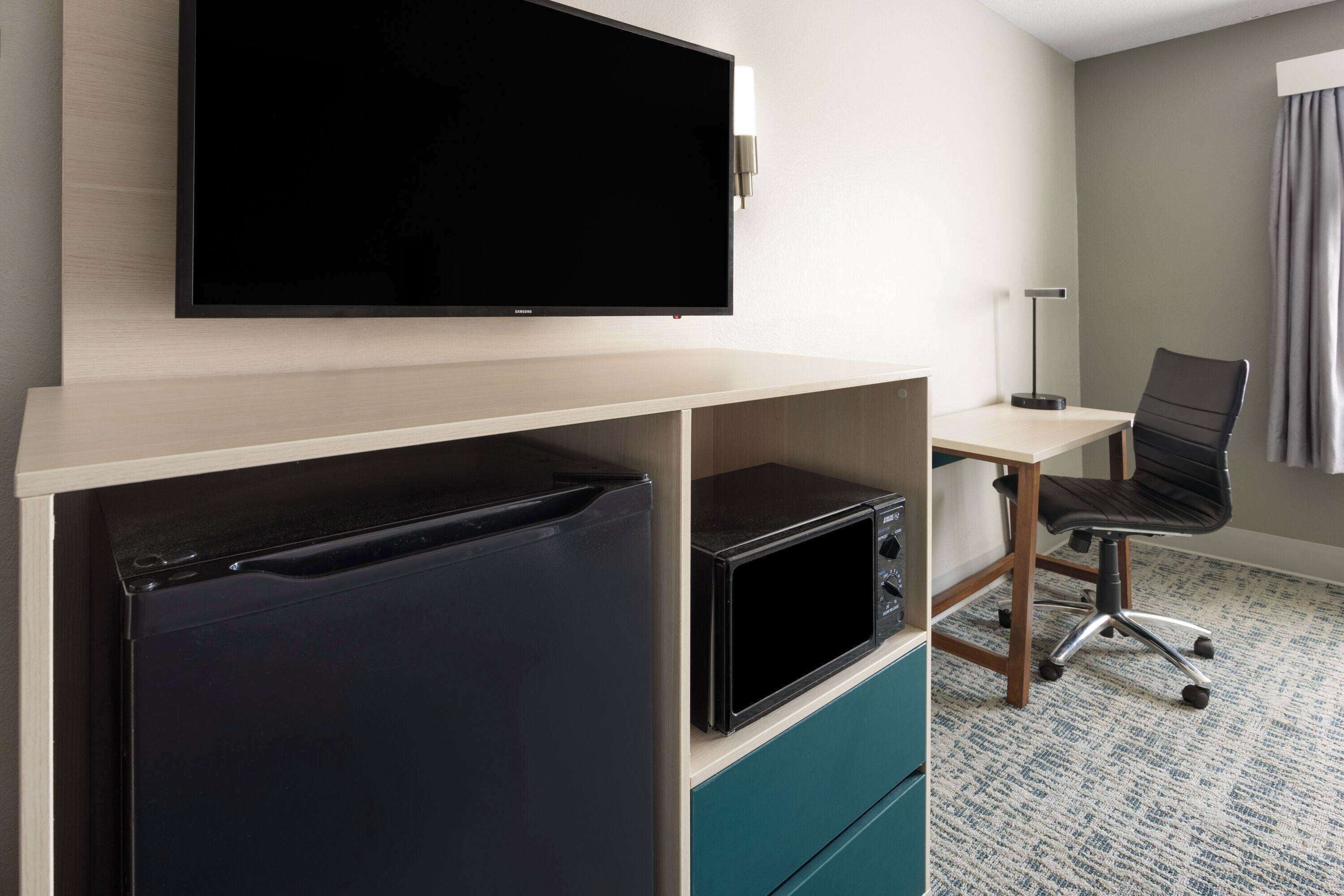 Habitación de hotel con televisión de pantalla plana en la pared, microondas, refrigerador pequeño, escritorio con silla y piso alfombrado.