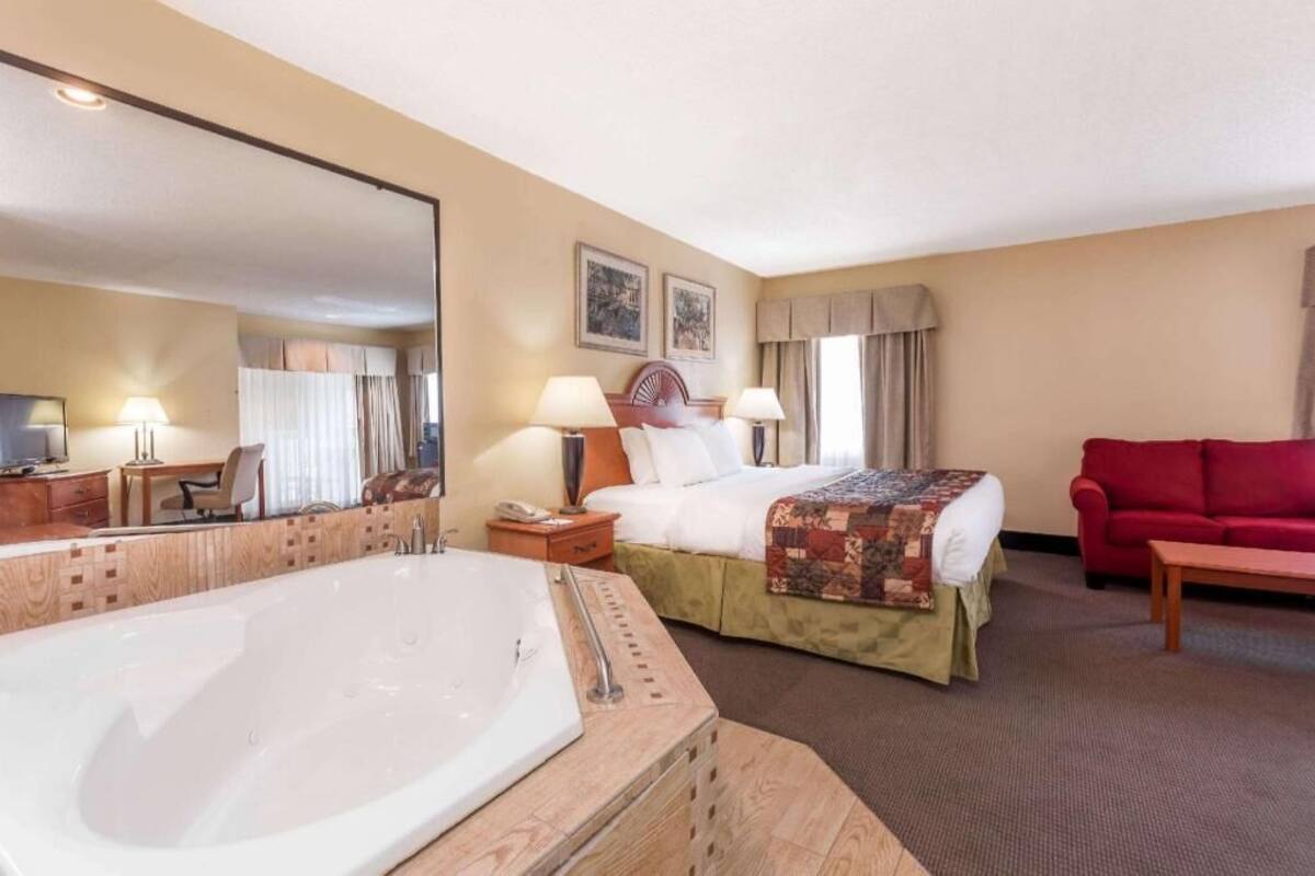 A hotel room with a large bathtub, a bed with a quilt, a red sofa, a desk with a lamp, and a mirror.