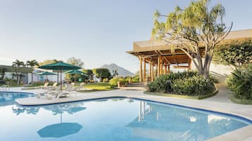 Una piscina con sillones de descanso y sombrillas, rodeada de vegetación y una pérgola de madera.