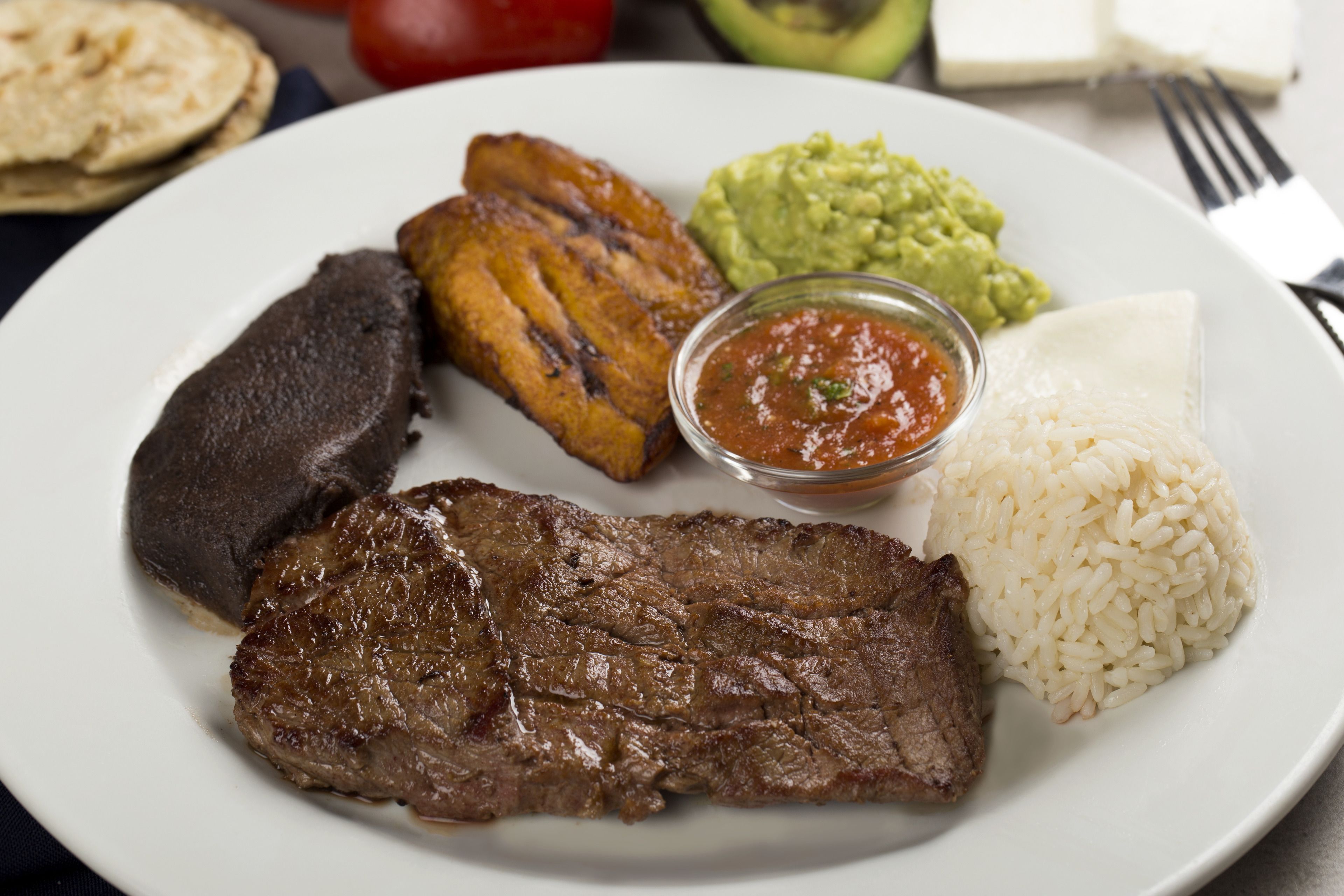 Un plato con bistec, frijoles negros, tostones, arroz y guacamole.