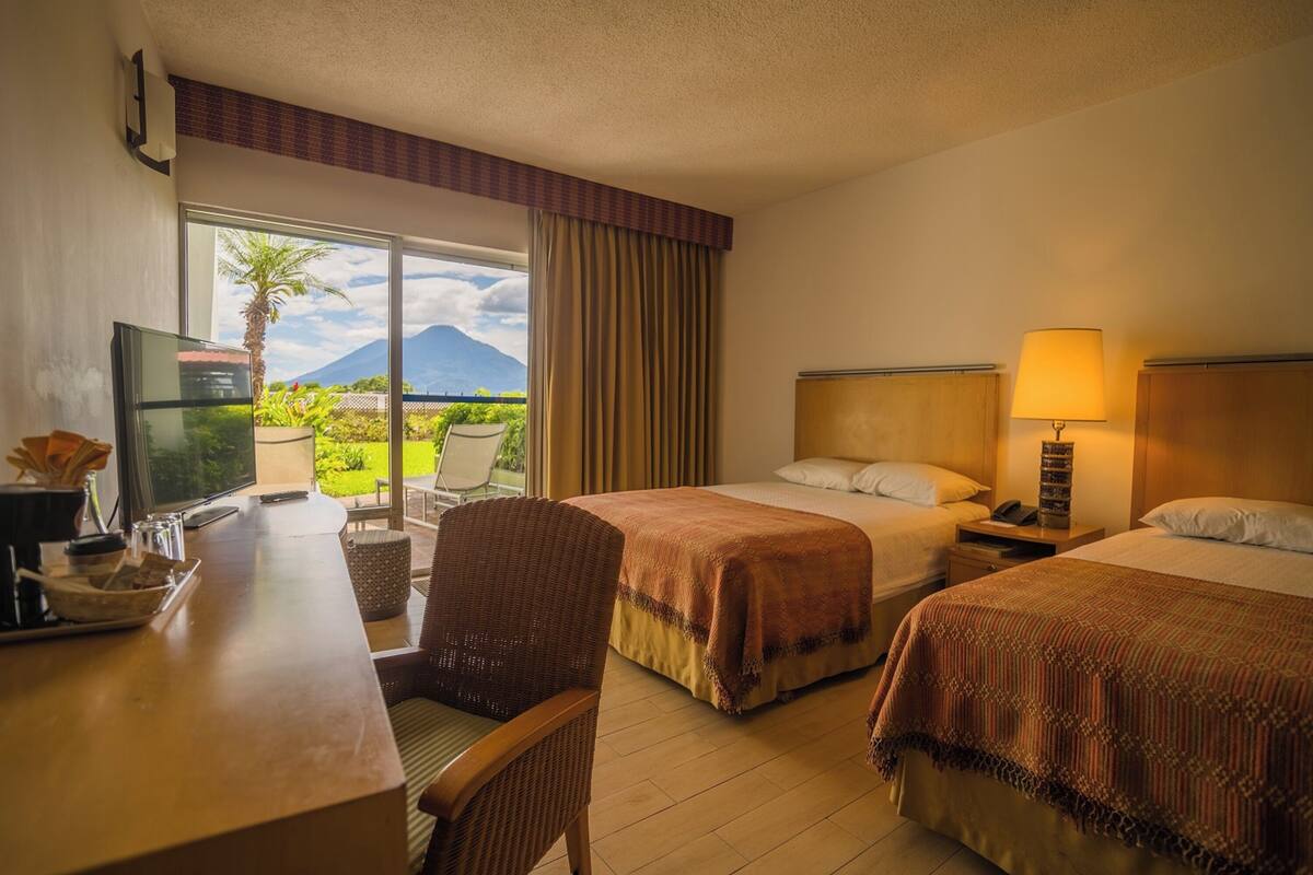 A hotel room with two beds, a desk with a television, and a view of a mountain.